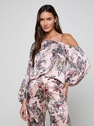 A woman poses with one hand on her hip against a plain background, wearing the Asami Chain One-Shoulder Blouse in a pink butterfly jungle print satin, styled with matching wide-leg pants.