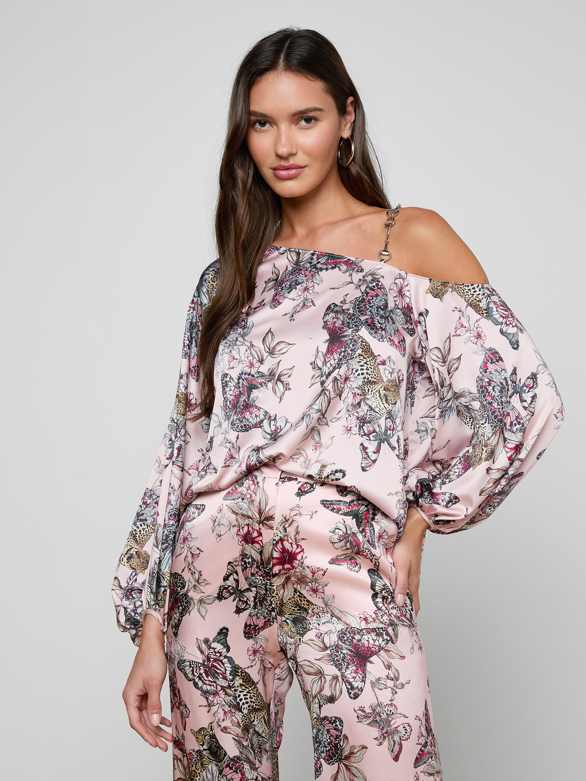 A woman poses with one hand on her hip against a plain background, wearing the Asami Chain One-Shoulder Blouse in a pink butterfly jungle print satin, styled with matching wide-leg pants.