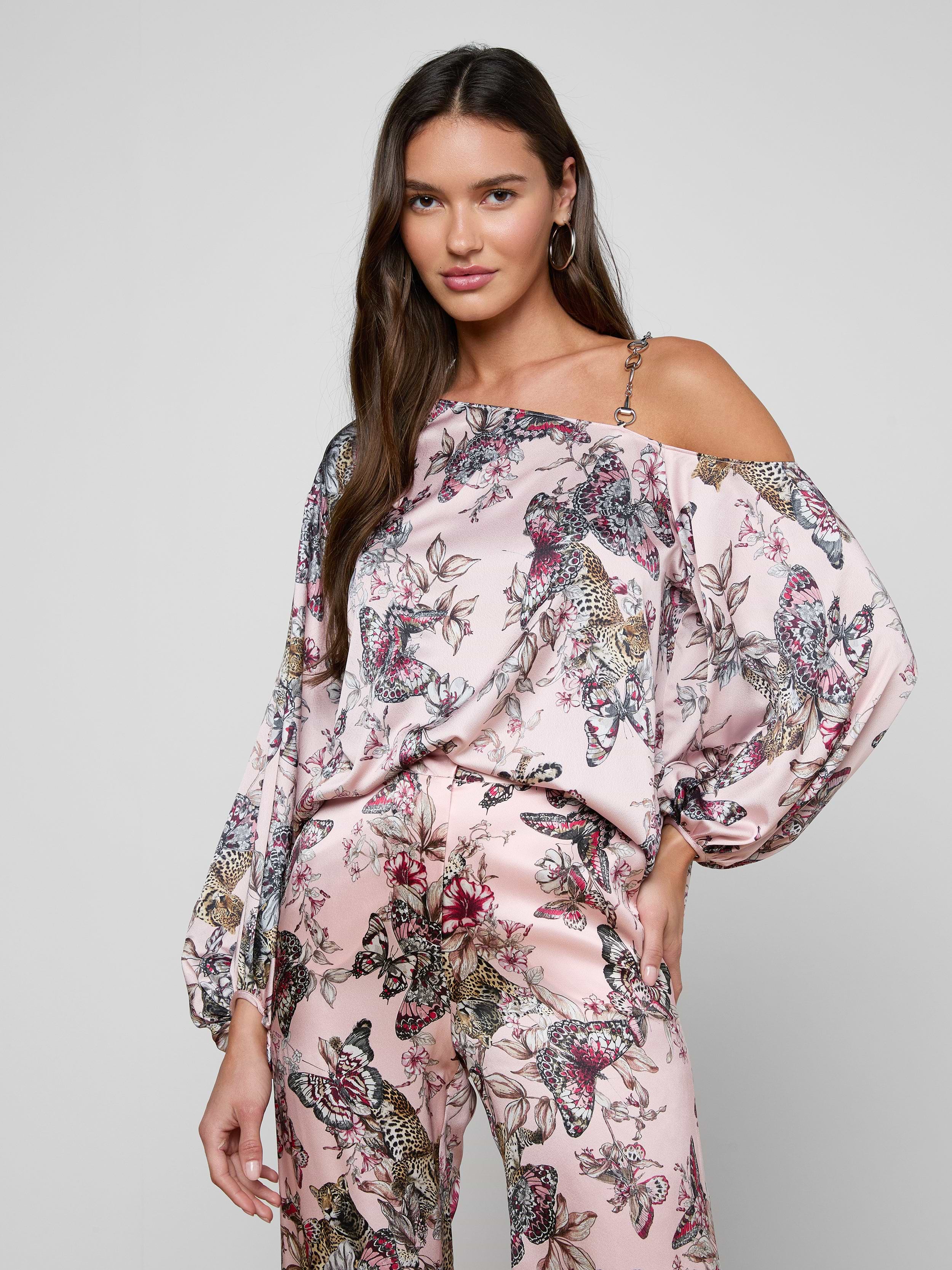 A woman poses with one hand on her hip against a plain background, wearing the Asami Chain One-Shoulder Blouse in a pink butterfly jungle print satin, styled with matching wide-leg pants.
