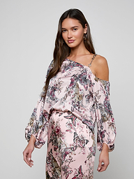 A woman wearing the Asami Chain One-Shoulder Blouse in light pink floral with puffy sleeves and matching pants stands against a plain background, looking at the camera with a relaxed expression.