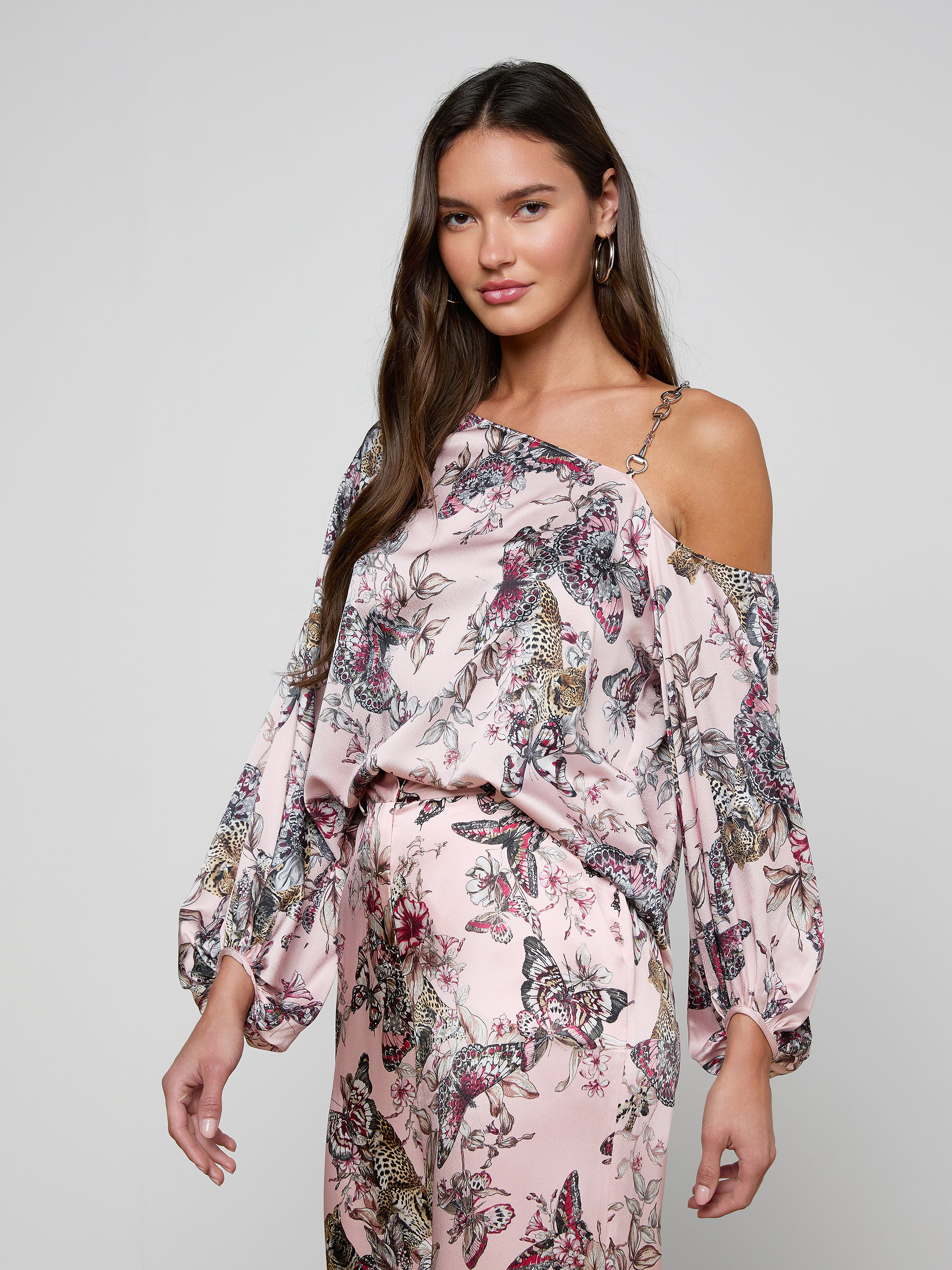 A woman wearing the Asami Chain One-Shoulder Blouse in light pink floral with puffy sleeves and matching pants stands against a plain background, looking at the camera with a relaxed expression.
