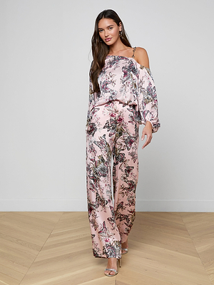 A woman with long brown hair wears the Asami Chain One-Shoulder Blouse and matching wide-leg pants, paired with metallic heels, standing on a light wood floor against a plain white wall.