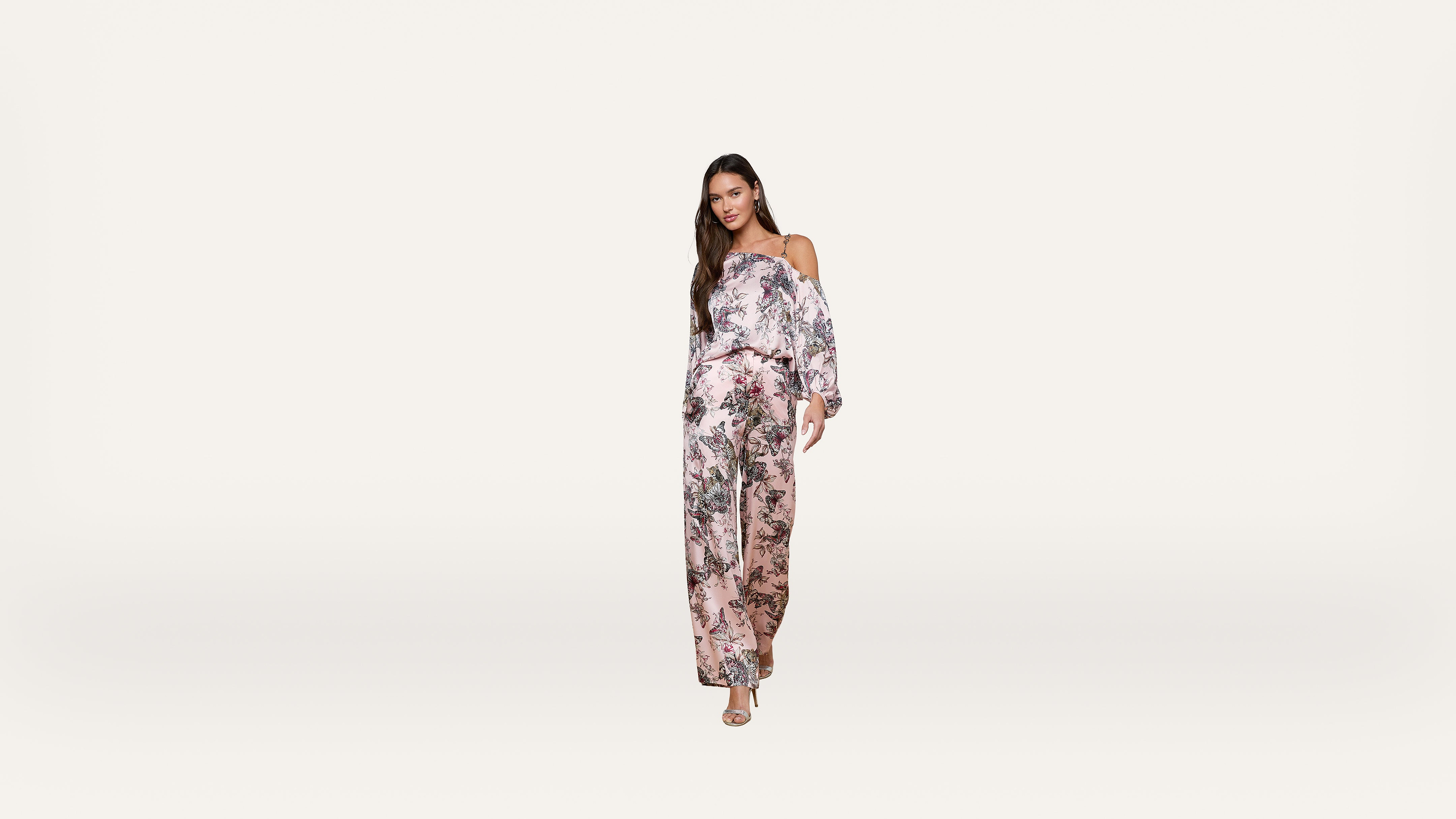Woman wearing a pink floral off-shoulder jumpsuit and heels stands against a plain white background, facing forward with a relaxed expression and long hair down.