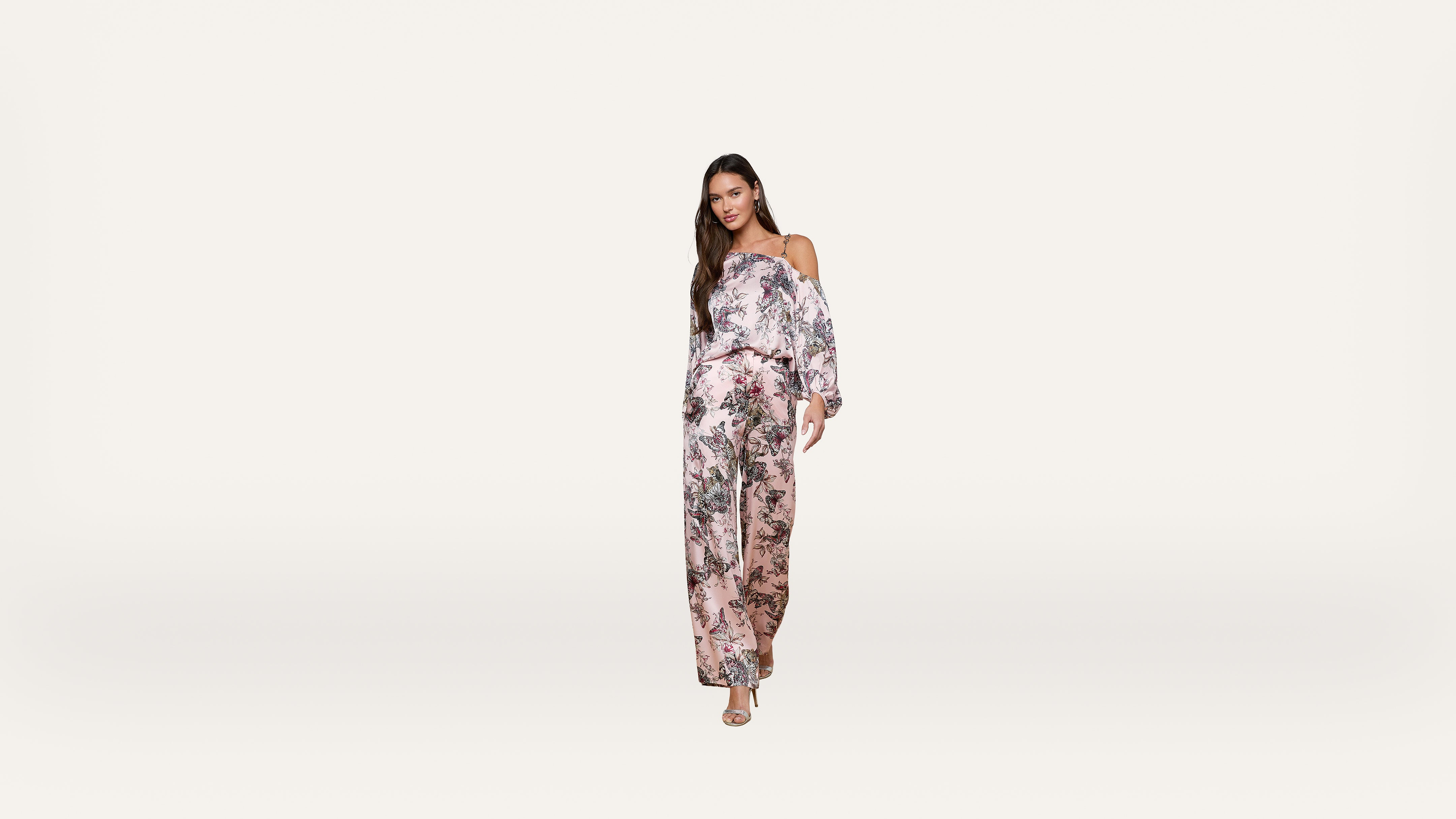 Woman wearing a pink floral off-shoulder jumpsuit and heels stands against a plain white background, facing forward with a relaxed expression and long hair down.
