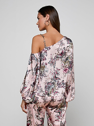 A woman with brown hair worn down and hoop earrings stands with her back to the camera, modeling the Asami Chain One-Shoulder Blouse in pink floral print with matching pants against a plain, light-colored background.