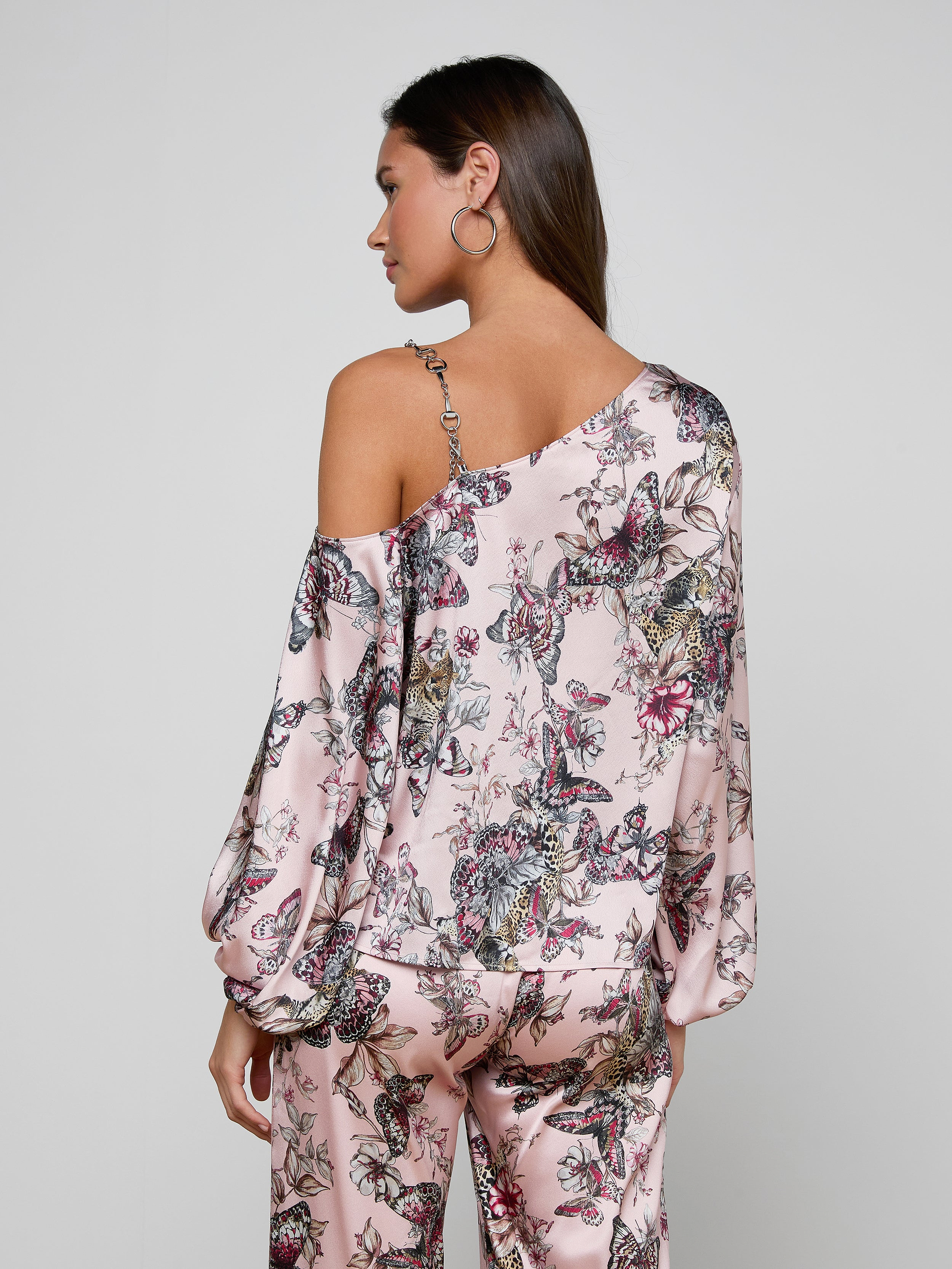 A woman with brown hair worn down and hoop earrings stands with her back to the camera, modeling the Asami Chain One-Shoulder Blouse in pink floral print with matching pants against a plain, light-colored background.