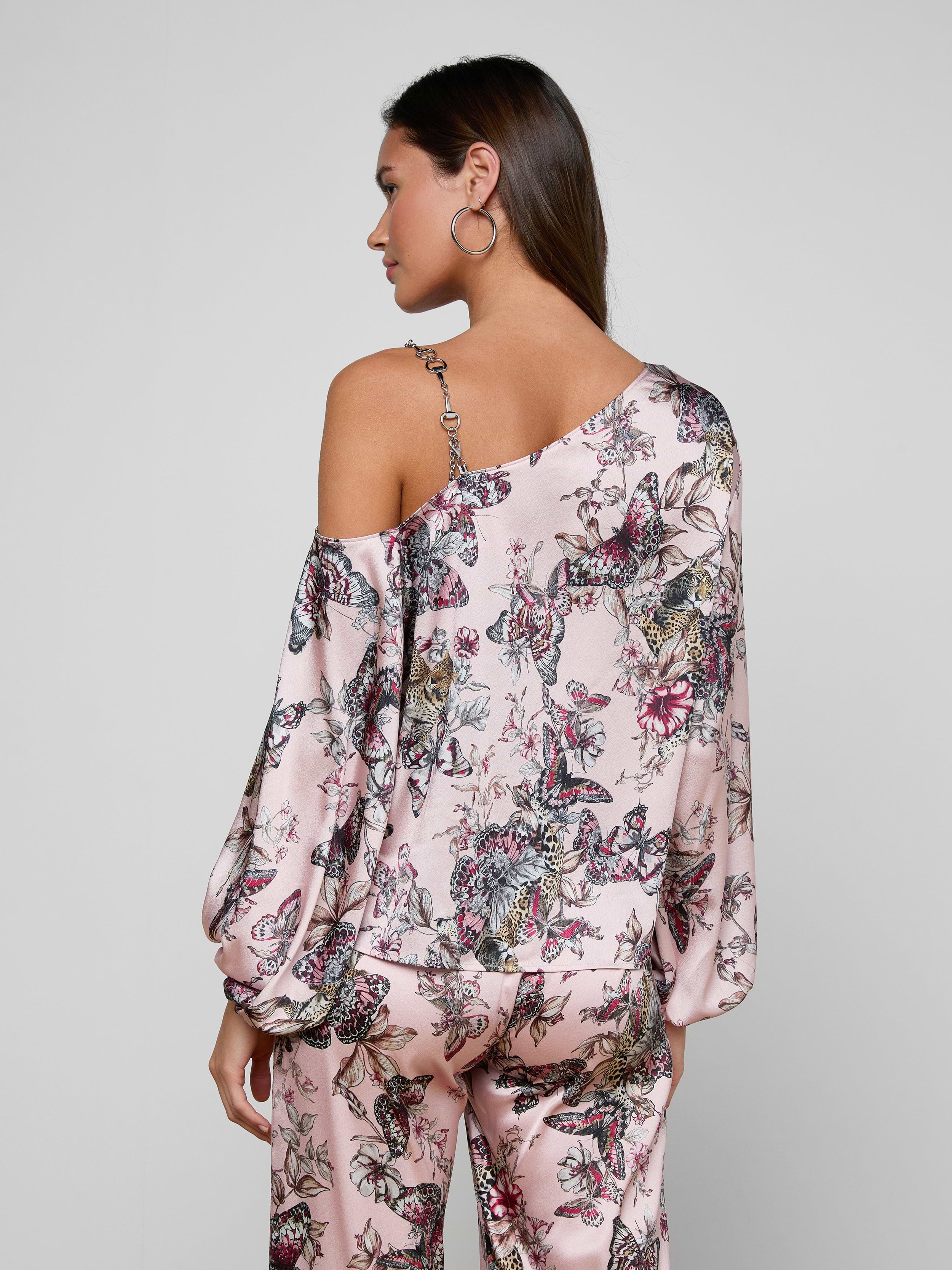 A woman with brown hair worn down and hoop earrings stands with her back to the camera, modeling the Asami Chain One-Shoulder Blouse in pink floral print with matching pants against a plain, light-colored background.