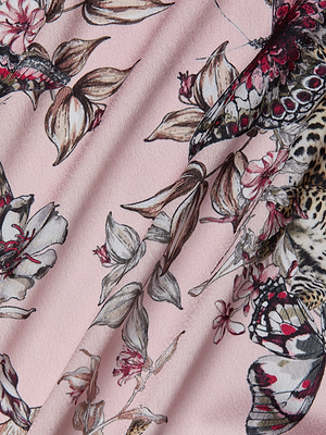 A close-up of the Asami Chain One-Shoulder Blouse shows pink fabric with a detailed butterfly jungle print, featuring black, white, red, and beige floral and butterfly motifs. Soft folds create gentle shadows and texture.