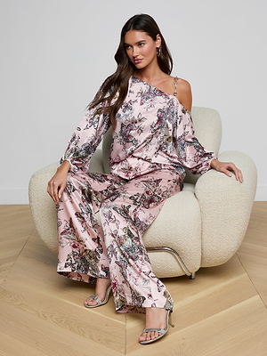 A woman with long brown hair sits on a cream armchair wearing the Asami Chain One-Shoulder Blouse in silky light pink with a butterfly jungle print, paired with matching wide-leg pants and silver heeled sandals, gazing to the side.