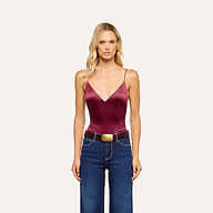 A woman with long blonde hair wears the Dara Stretch-Satin Bodysuit in maroon, featuring adjustable straps. She pairs it with blue jeans, a brown belt with a large buckle, and gold earrings, standing facing forward against a plain light background.