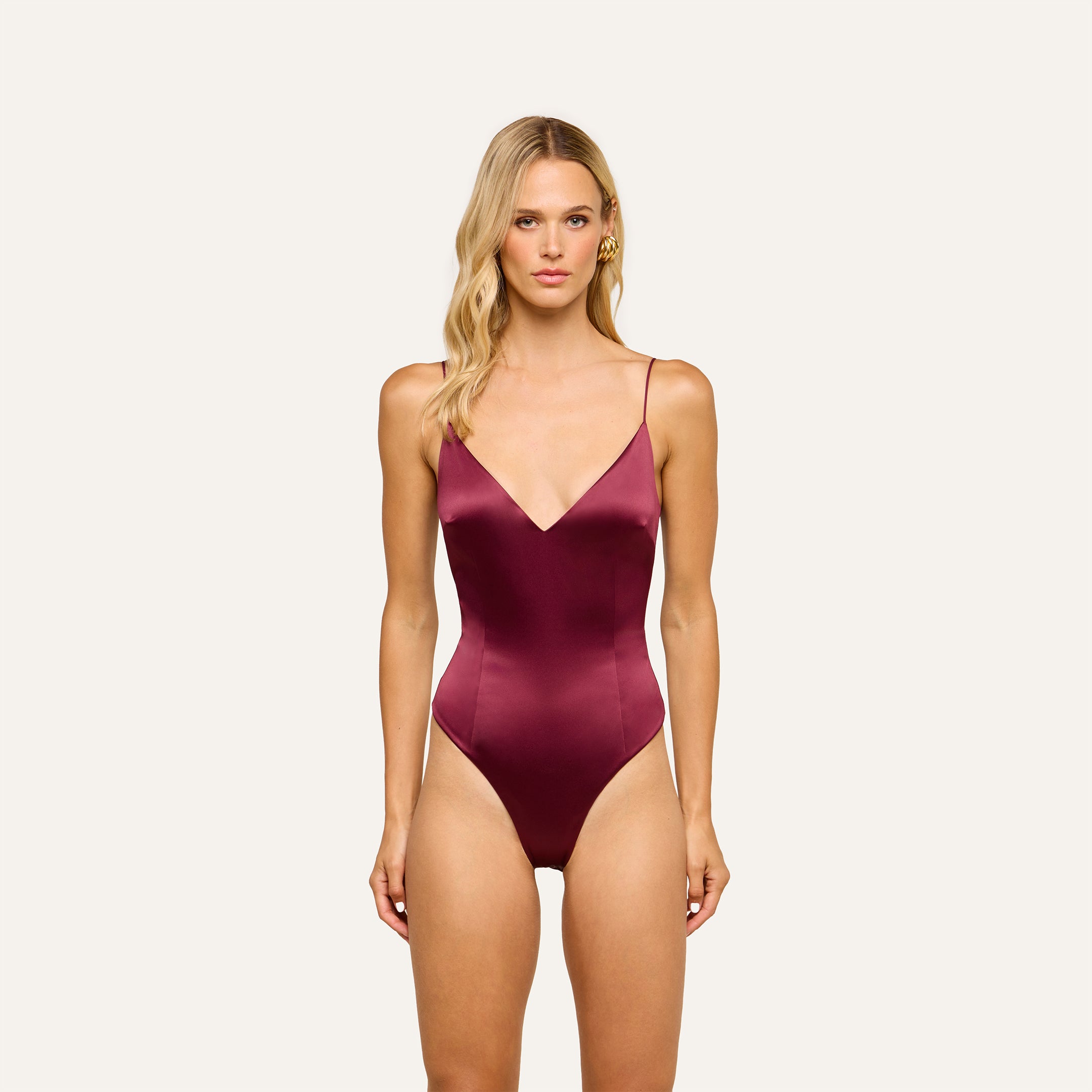 A woman wearing the Dara Stretch-Satin Bodysuit in deep burgundy, featuring adjustable straps, stands against a plain, light background, facing forward.