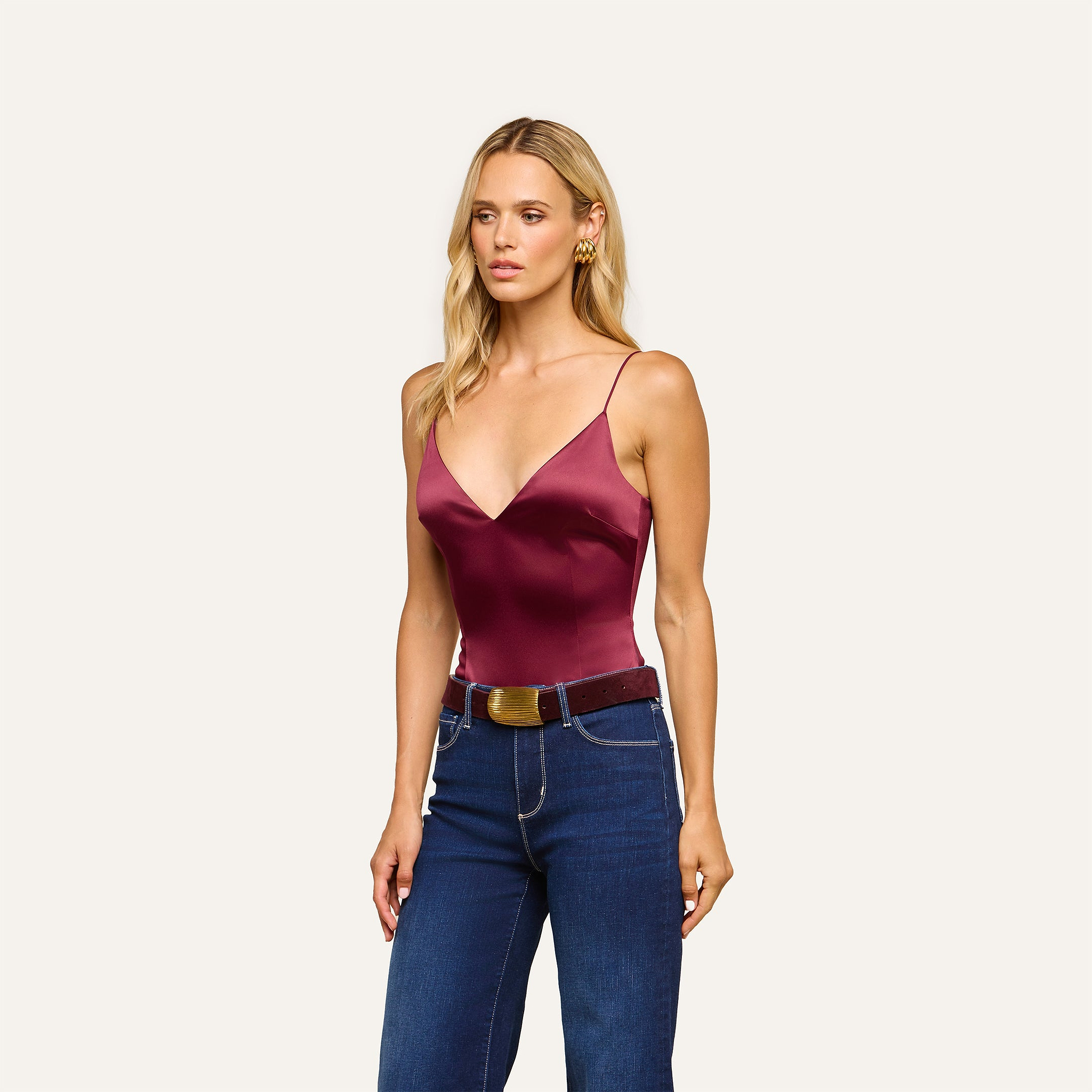 A woman with long blonde hair wears the Dara Stretch-Satin Bodysuit in burgundy, which features adjustable straps, paired with blue high-waisted jeans and a brown belt, standing against a plain light background.
