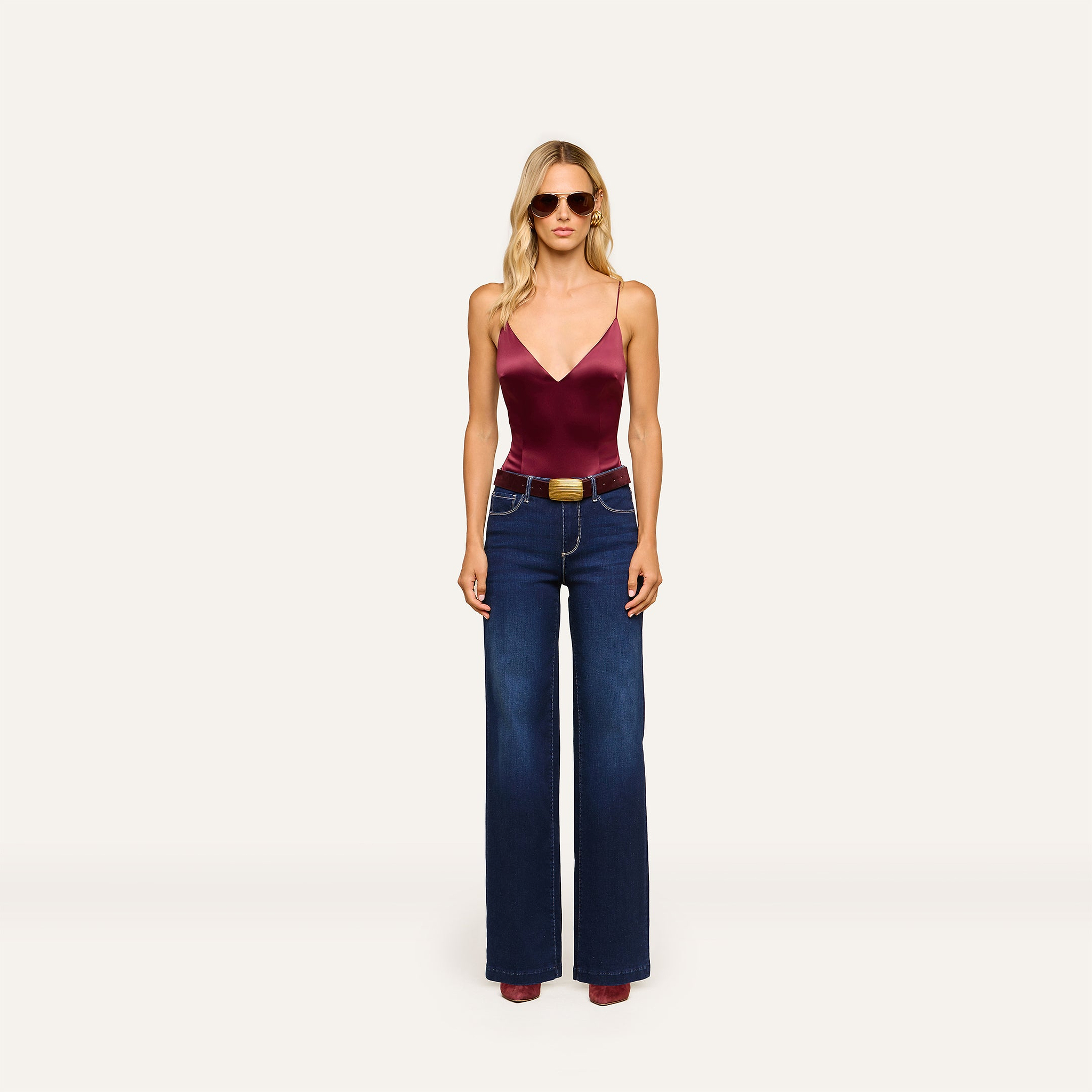 Wearing sunglasses and the Dara Stretch-Satin Bodysuit with adjustable straps, styled with wide-leg blue jeans, a brown belt, and burgundy shoes, the woman stands against a plain white background.