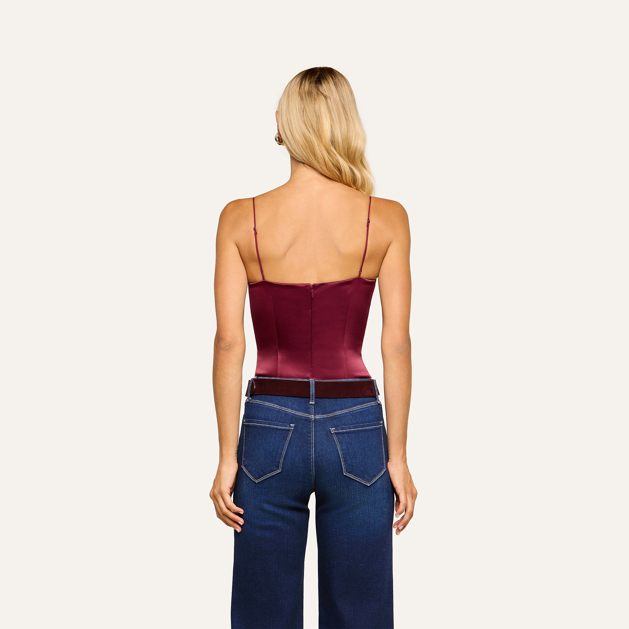 A woman with long blonde hair is facing away, wearing the Dara Stretch-Satin Bodysuit in burgundy, which features adjustable straps, paired with high-waisted dark blue jeans. The background is plain and light-colored.