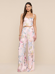 Une femme aux longs cheveux bruns porte le bustier Sonya en rose pâle, associé à un pantalon large assorti à limprimé feuille de palmier coloré. Elle porte des boucles doreilles et un bracelet en or sur fond beige.