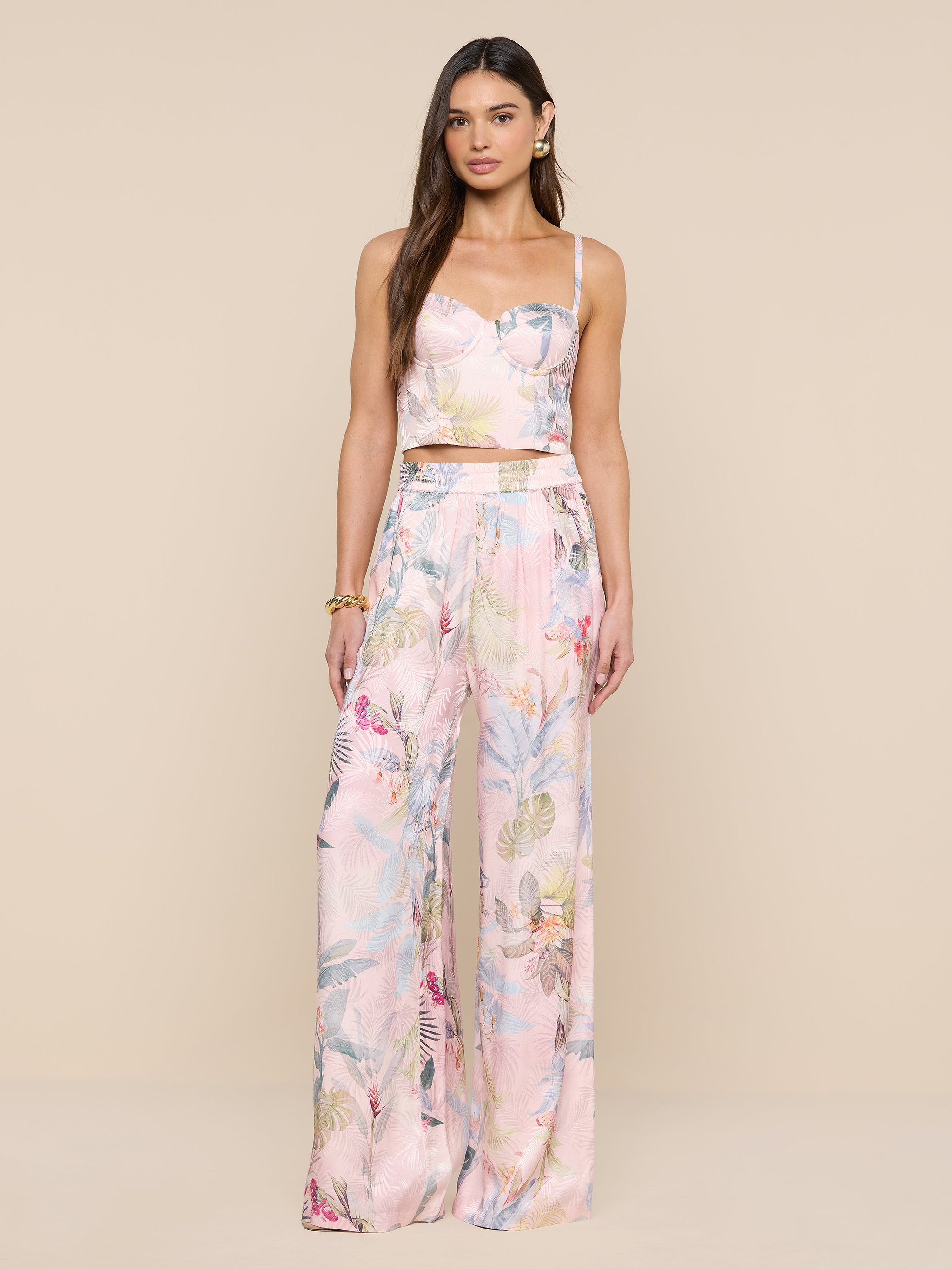Une femme aux longs cheveux bruns porte le bustier Sonya en rose pâle, associé à un pantalon large assorti à limprimé feuille de palmier coloré. Elle porte des boucles doreilles et un bracelet en or sur fond beige.