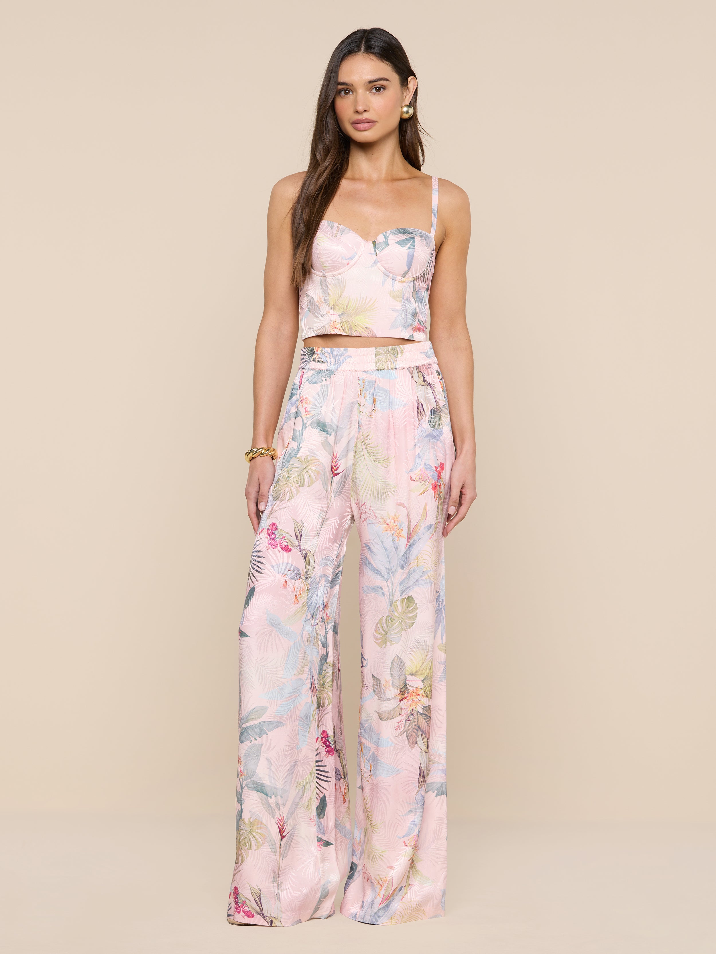 Une femme aux longs cheveux bruns porte le bustier Sonya en rose pâle, associé à un pantalon large assorti à limprimé feuille de palmier coloré. Elle porte des boucles doreilles et un bracelet en or sur fond beige.