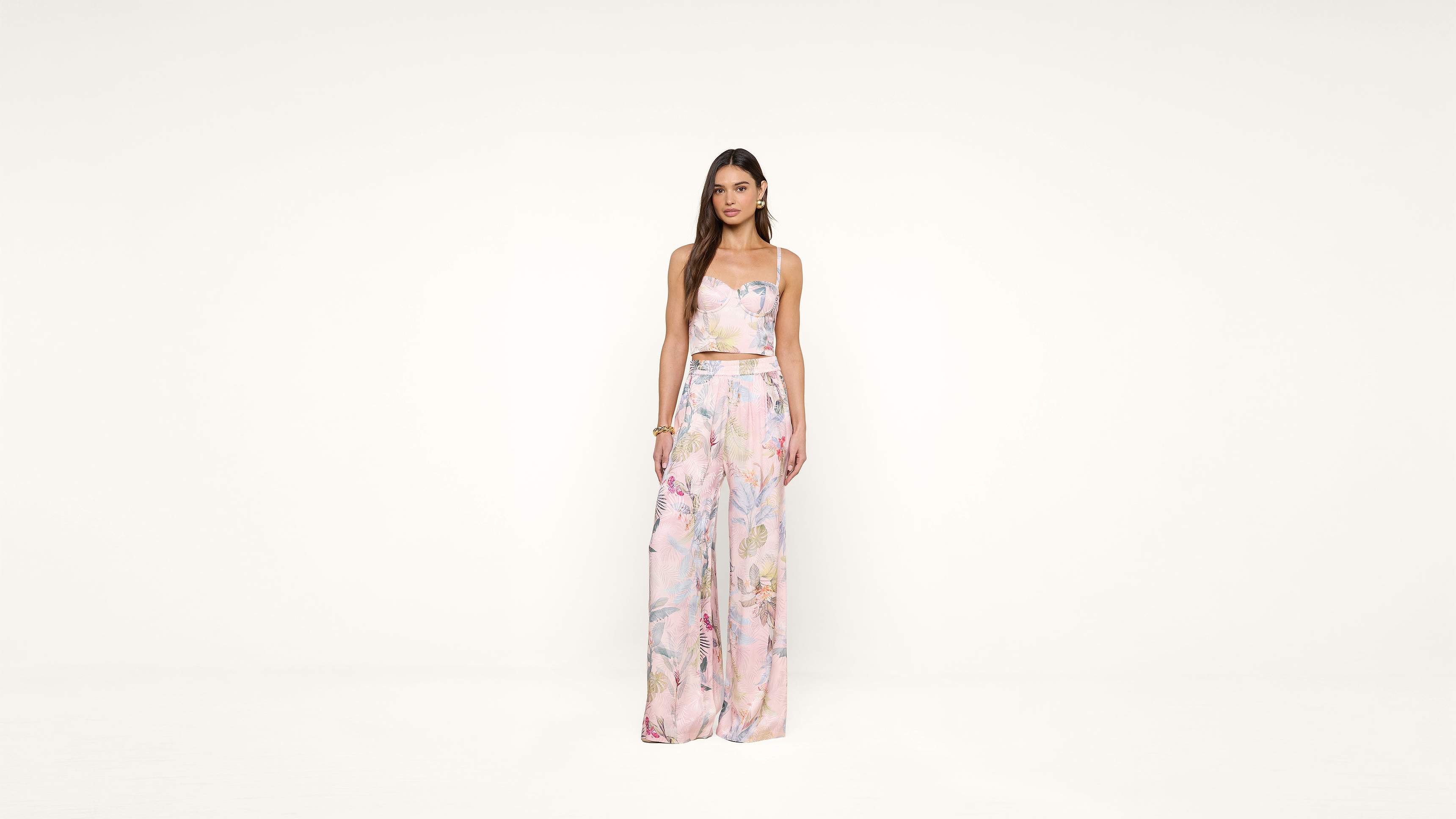 A woman stands against a plain white background, wearing a sleeveless, pastel floral crop top and matching wide-leg pants. She has long, straight hair and a neutral expression.