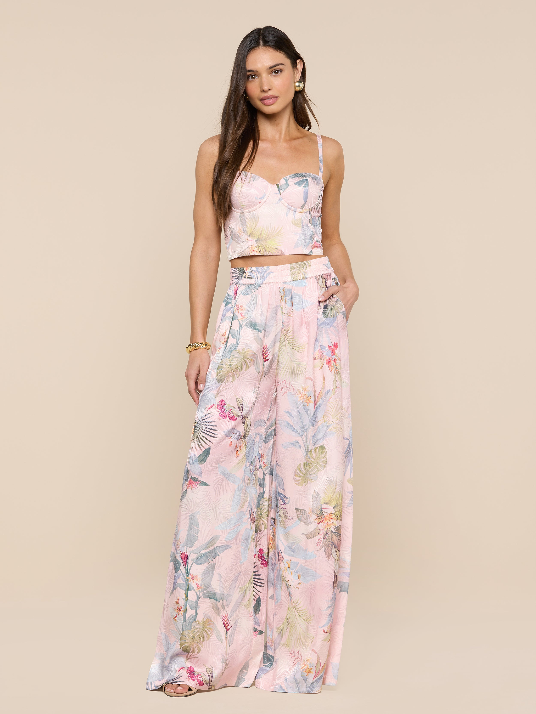 Une femme aux longs cheveux bruns se tient sur un fond beige, portant le Sonya Bustier dans un ensemble deux pièces imprimé de feuilles de palmier roses, composé dun bustier ajusté et dun pantalon large à taille haute, une main dans la poche et une expression neutre.