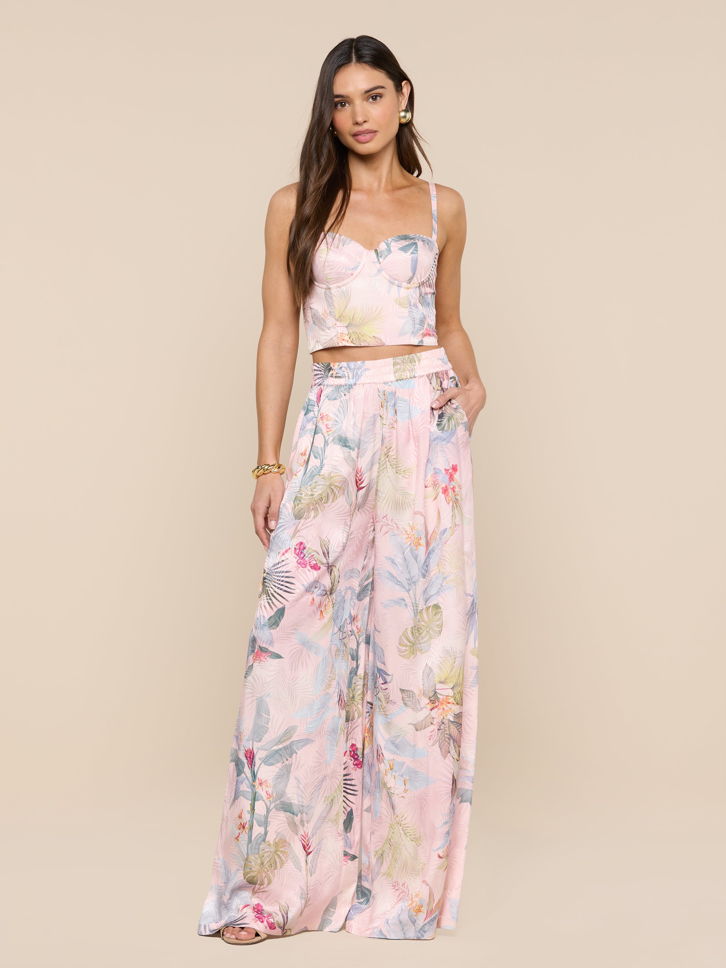 Une femme aux longs cheveux bruns se tient sur un fond beige, portant le Sonya Bustier dans un ensemble deux pièces imprimé de feuilles de palmier roses, composé dun bustier ajusté et dun pantalon large à taille haute, une main dans la poche et une expression neutre.