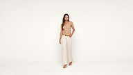A woman stands against a plain white background, wearing a beige lace camisole, cream-colored wide-leg pants with a gold belt, and beige sandals. She has long hair and a relaxed expression.