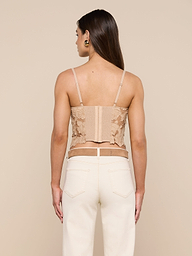 A woman with long brown hair, wearing the Sonya Bustier—a beige cotton eyelet top with adjustable straps—and high-waisted white jeans, stands facing away from the camera against a plain off-white background.