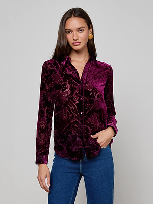 A woman with long dark hair wears the L'AGENCE Laurent Velvet Blouse, a relaxed fit, long-sleeve purple burnout velvet shirt with a floral pattern, paired with blue jeans. She stands against a light gray background, one hand in her pocket.