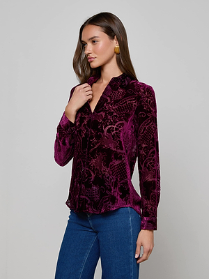 A woman wears the L'AGENCE Laurent Velvet Blouse in deep purple with a subtle pattern and relaxed fit, paired with blue jeans, standing against a gray background looking to the side and touching her collar.