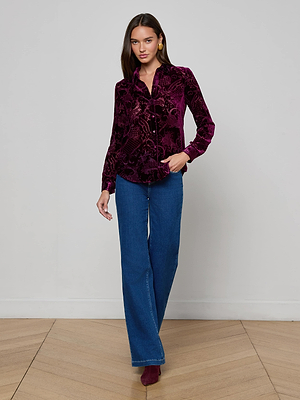 A woman poses indoors against a light backdrop wearing the L'AGENCE Laurent Velvet Blouse, styled with blue wide-leg jeans and purple heeled shoes. One hand is in her pocket, showcasing the blouse's relaxed fit.