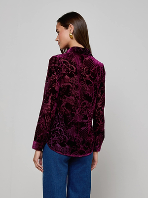 A woman with long brown hair faces away, showcasing the relaxed fit and long sleeves of the deep purple Laurent Velvet Blouse by L'AGENCE, paired with blue jeans and gold earrings. The background is plain and light-colored.