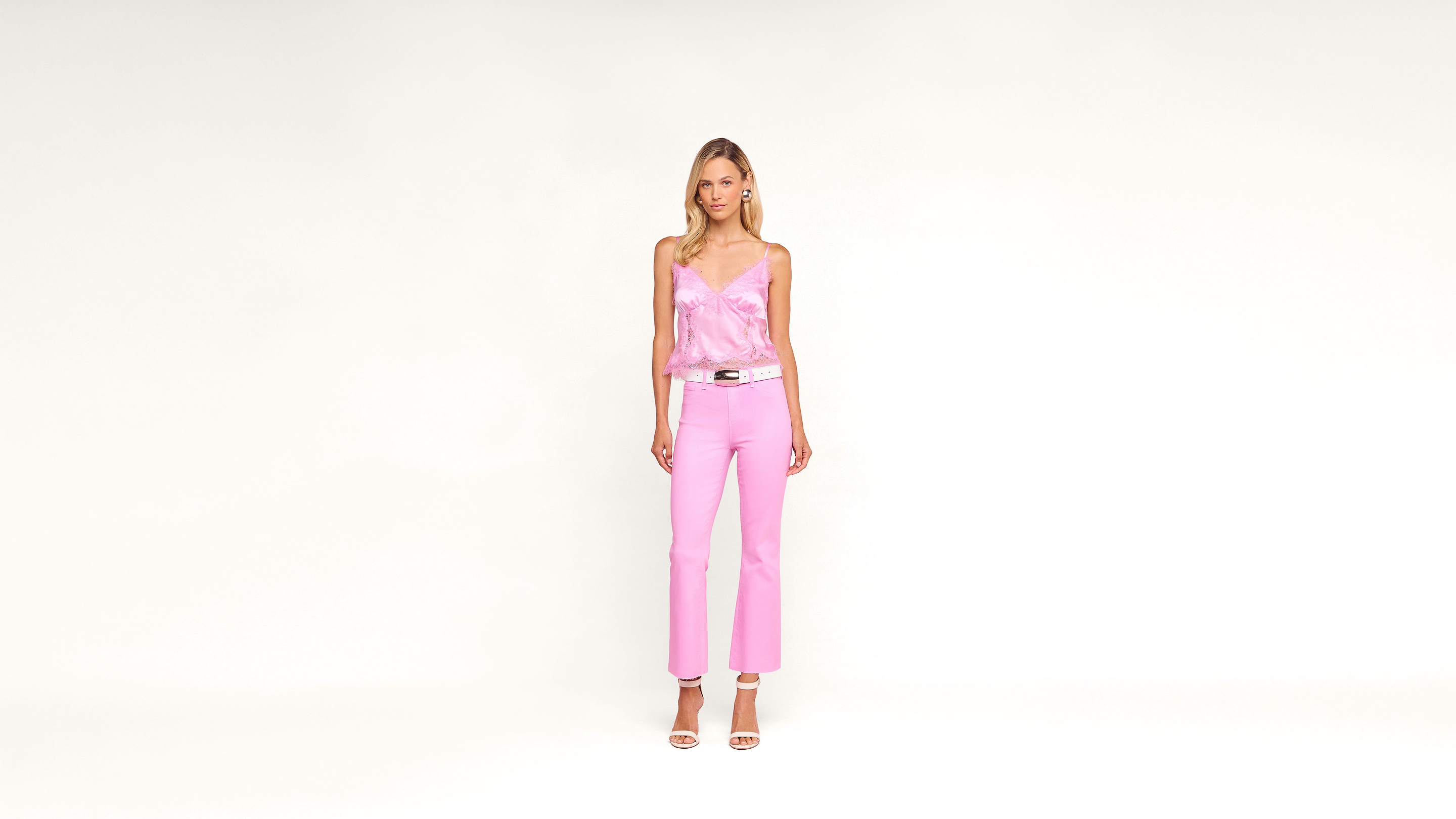 Woman stands against a plain white background, wearing a light pink satin camisole, pink pants, white belt, and white high-heeled sandals. She has long hair and a neutral expression.
