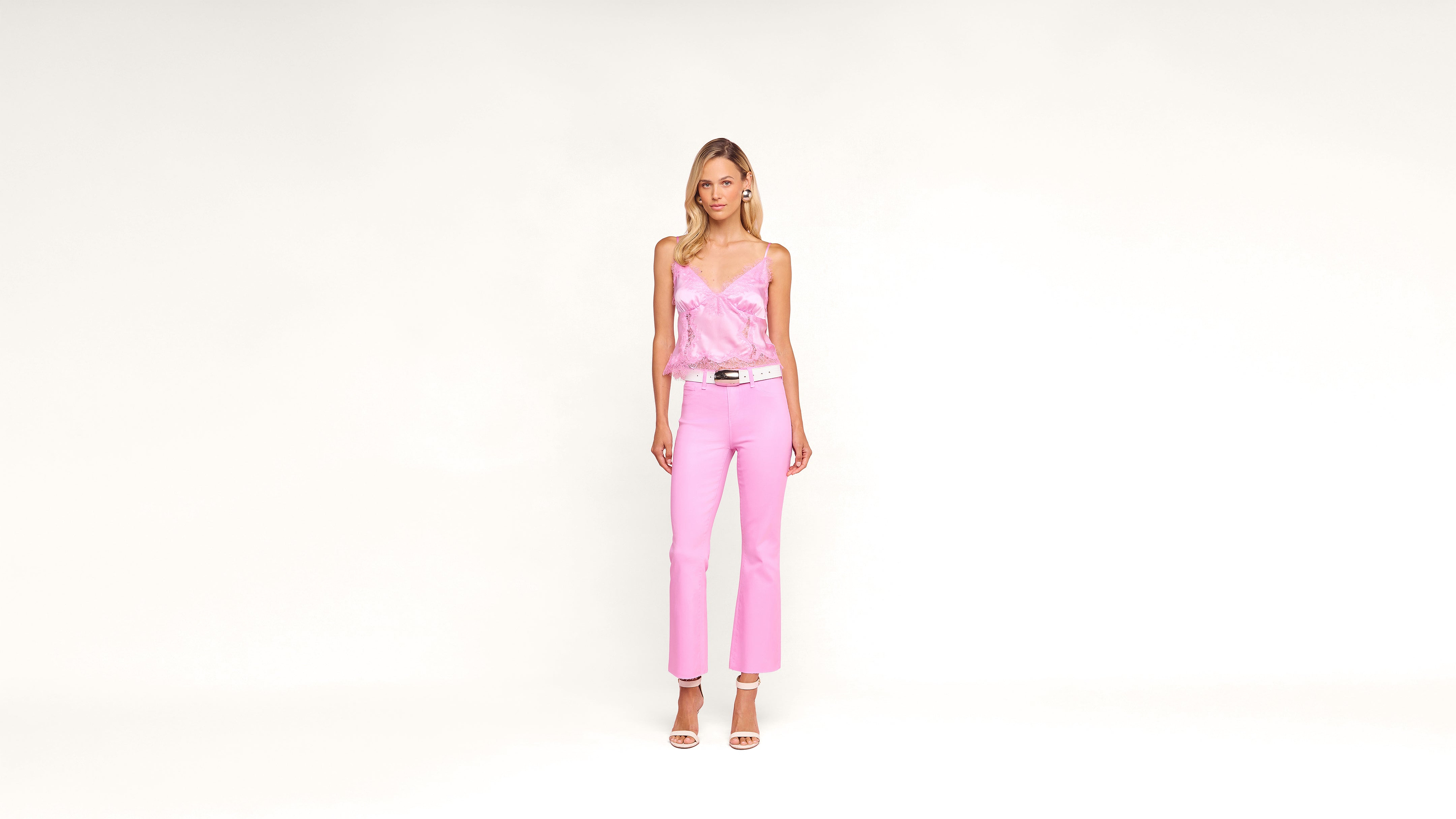 Woman stands against a plain white background, wearing a light pink satin camisole, pink pants, white belt, and white high-heeled sandals. She has long hair and a neutral expression.