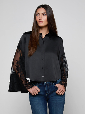 A woman wears the L'AGENCE Capri Lace Sleeve Blouse—a relaxed fit button-down with sheer lace bell sleeves—and blue jeans, standing with hands on hips and looking to the side against a plain gray background.