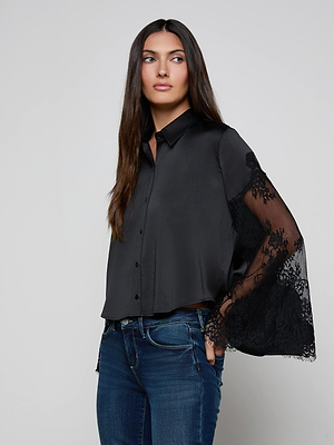 A woman with long brown hair wears the L'AGENCE Capri Lace Sleeve Blouse, a relaxed-fit black satin button-down with sheer lace bell sleeves, paired with blue jeans against a plain light gray background.