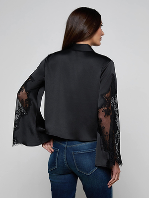 A woman with long brown hair, wearing the L'AGENCE Capri Lace Sleeve Blouse with sheer lace bell sleeves and blue jeans, stands facing away from the camera with one hand on her hip.