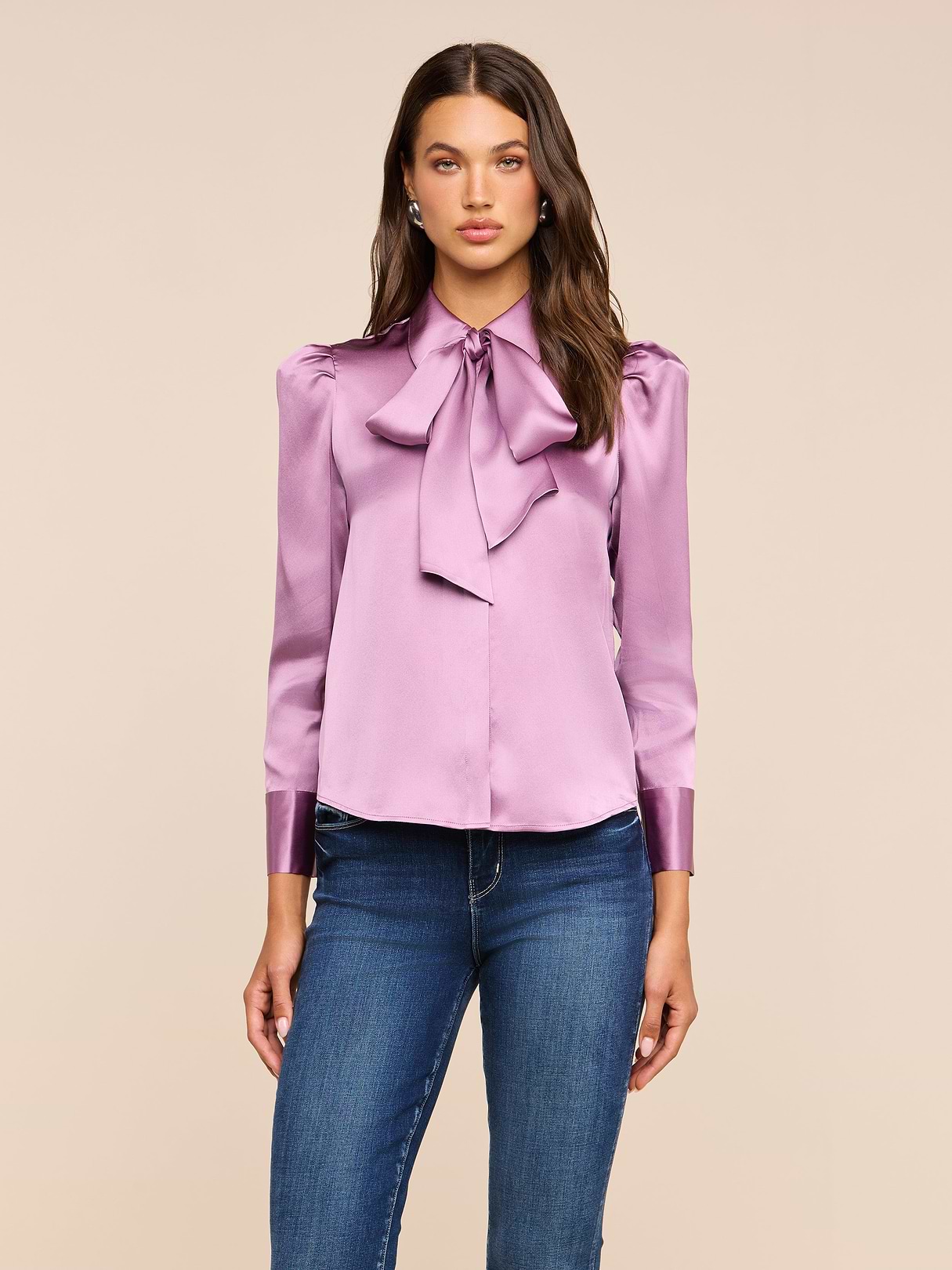 A woman stands against a plain background, wearing the Honey Silk Blouse—a long-sleeved silk charmeuse top with a large bow at the collar—paired with blue jeans.