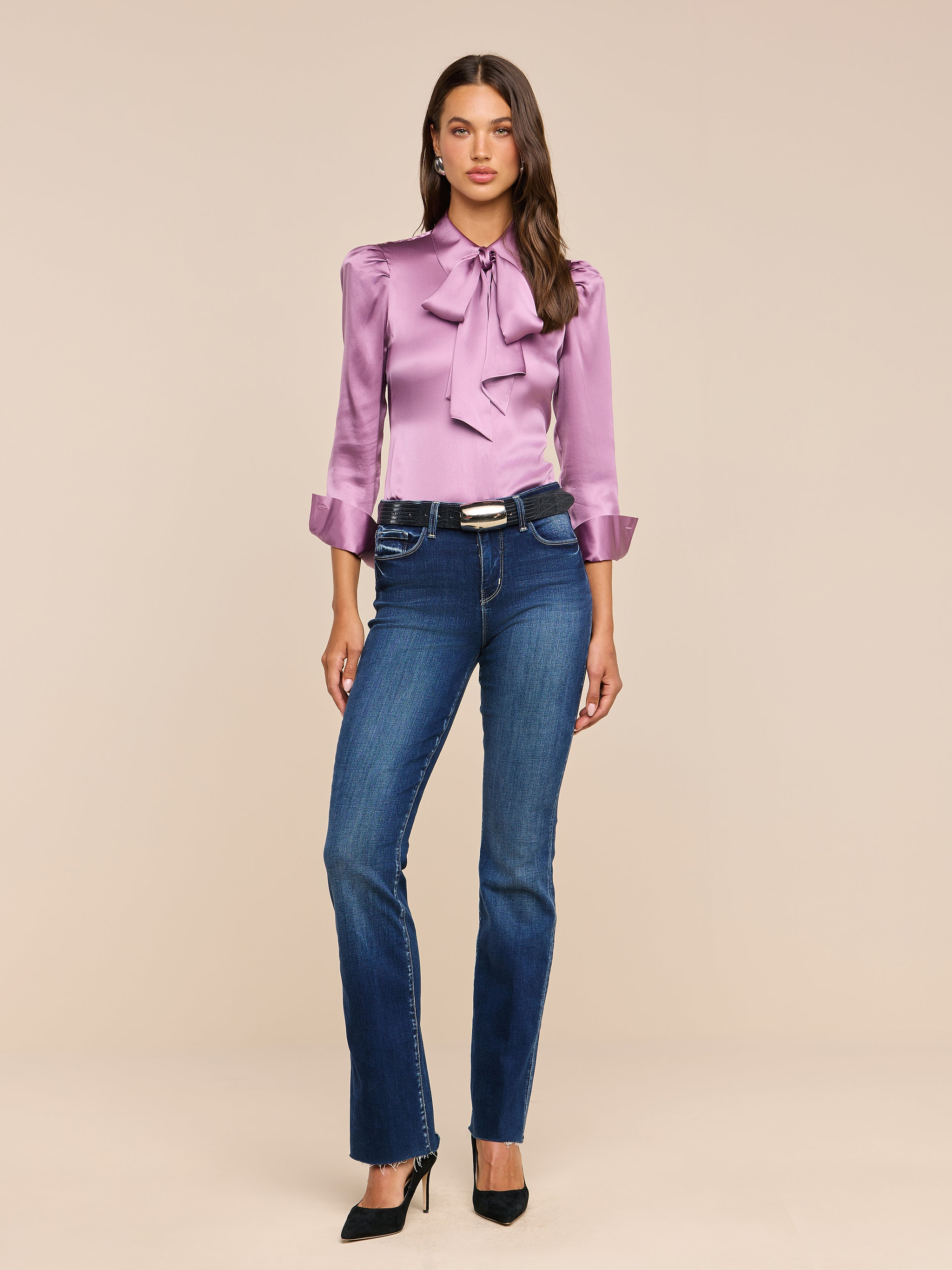A woman stands facing forward against a plain white background, wearing the Honey Silk Blouse—a shiny lavender silk top with a bow—paired with blue jeans, a black belt, and black heels.