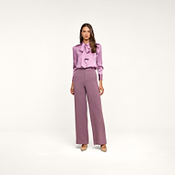 A woman with long hair stands against a plain white background wearing the Honey Silk Blouse in lavender, paired with high-waisted, wide-leg mauve trousers and nude heels.