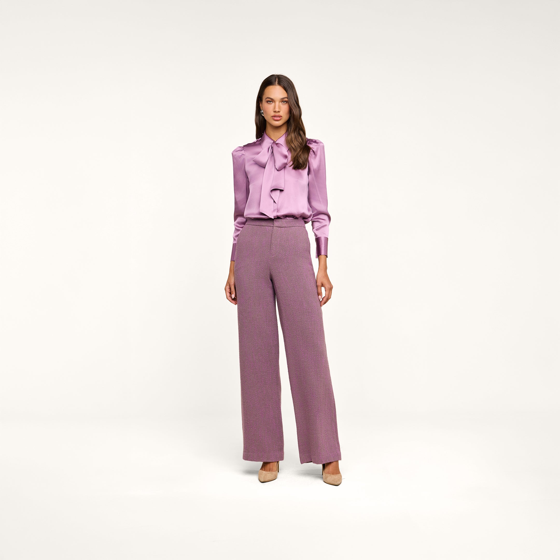 A woman with long hair stands against a plain white background wearing the Honey Silk Blouse in lavender, paired with high-waisted, wide-leg mauve trousers and nude heels.