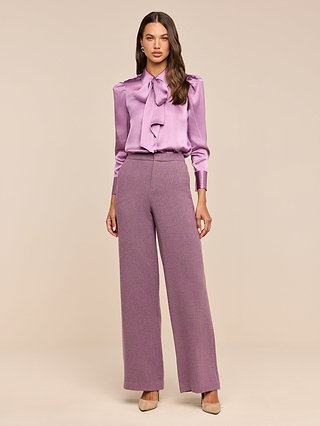 A woman stands in a studio wearing the Honey Silk Blouse—a shiny lavender silk charmeuse tie-neck top—paired with wide-leg mauve trousers and beige heels against a neutral background.