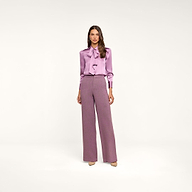 Woman standing against a plain background, wearing the Honey Silk Blouse in wisteria purple with a tie neck and long sleeves, paired with high-waisted, wide-leg mauve trousers and beige heels.