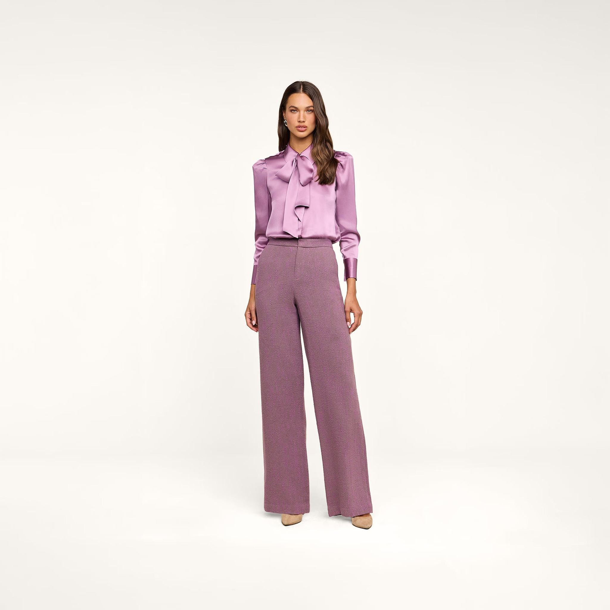 Woman standing against a plain background, wearing the Honey Silk Blouse in wisteria purple with a tie neck and long sleeves, paired with high-waisted, wide-leg mauve trousers and beige heels.