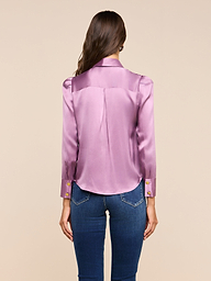 A woman with long brown hair, wearing the Honey Silk Blouse in a shiny lavender silk charmeuse paired with blue jeans, stands facing away from the camera against a plain light background.