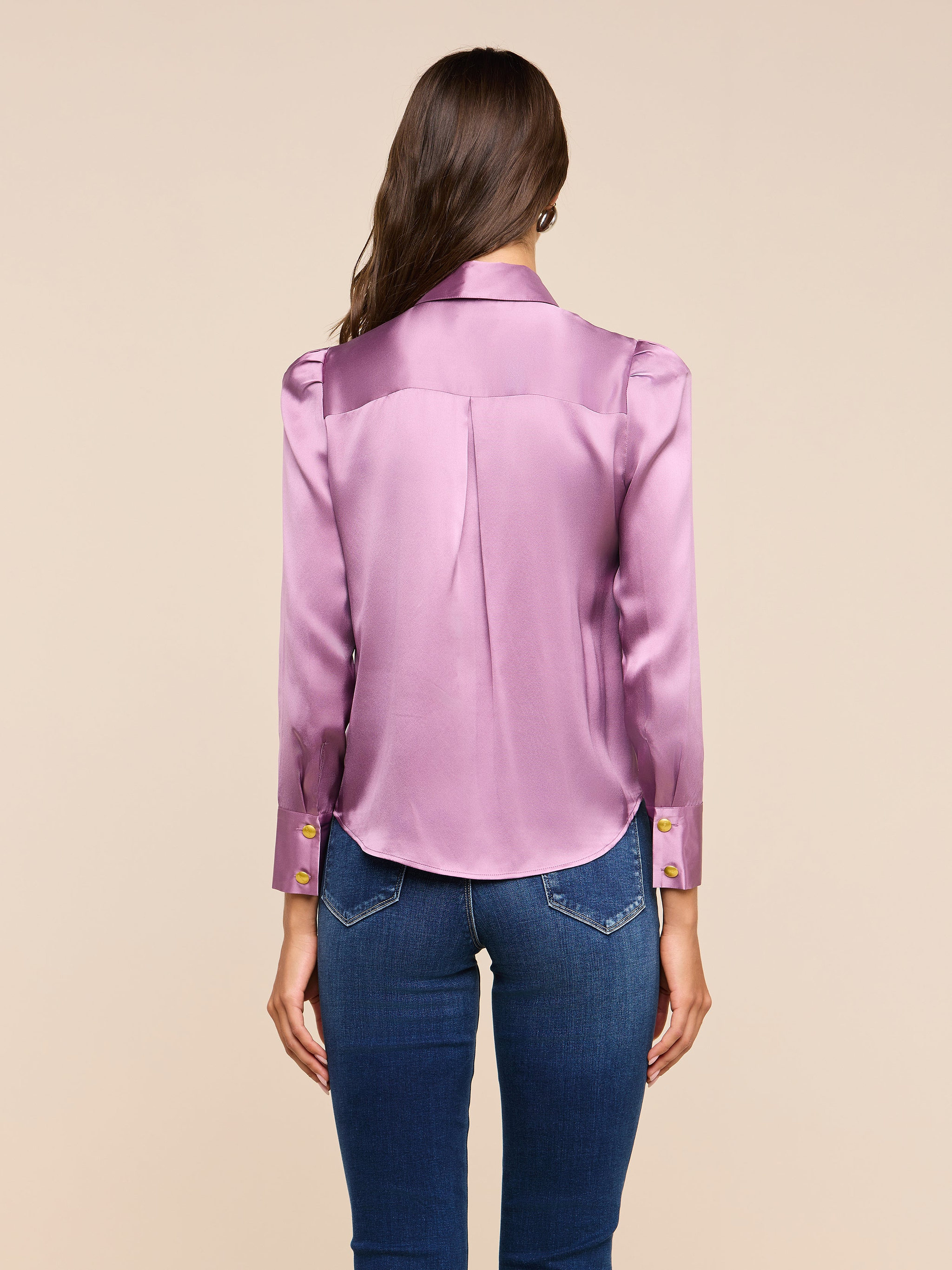 A woman with long brown hair, wearing the Honey Silk Blouse in a shiny lavender silk charmeuse paired with blue jeans, stands facing away from the camera against a plain light background.