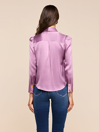 A woman stands facing away, wearing the Honey Silk Blouse—an elegant, shiny lavender long-sleeve top with gold buttons—paired with blue jeans, against a plain beige background.