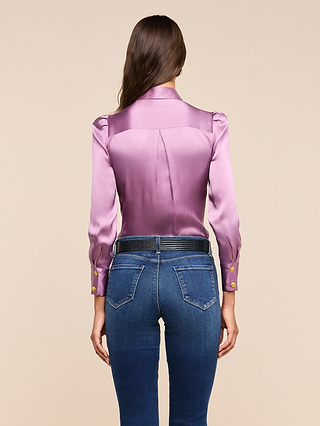 A woman stands facing away against a beige background, wearing the Honey Silk Blouse—a shiny lavender silk charmeuse top with gold buttons—paired with dark blue jeans.