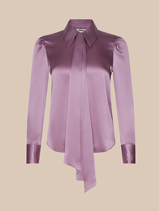 The Honey Silk Blouse is crafted from shiny lavender silk charmeuse and designed with long sleeves, a pointed collar, and a wide, extended necktie detail. This tie-neck blouse also features buttoned cuffs and a gently curved hem, shown against a plain light beige background.