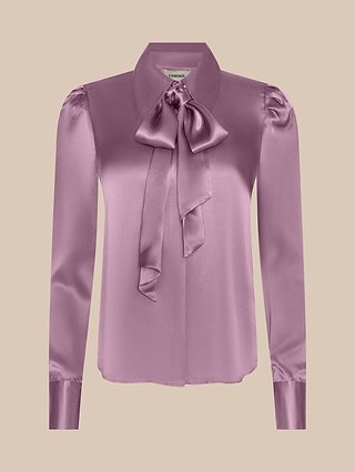 The Honey Silk Blouse is a long-sleeve, light purple silk charmeuse top featuring a large bow tie at the collar and buttoned cuffs, shown on a beige background.