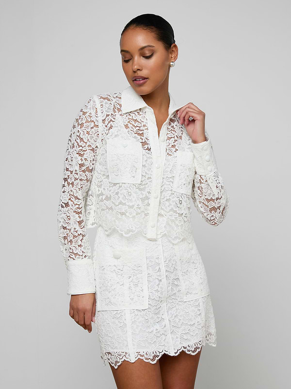 A woman wearing the Roelle Lace Blouse—a white, long-sleeve, floral lace top with patch pockets—stands against a plain light background, looking to the side with one hand raised near her face.