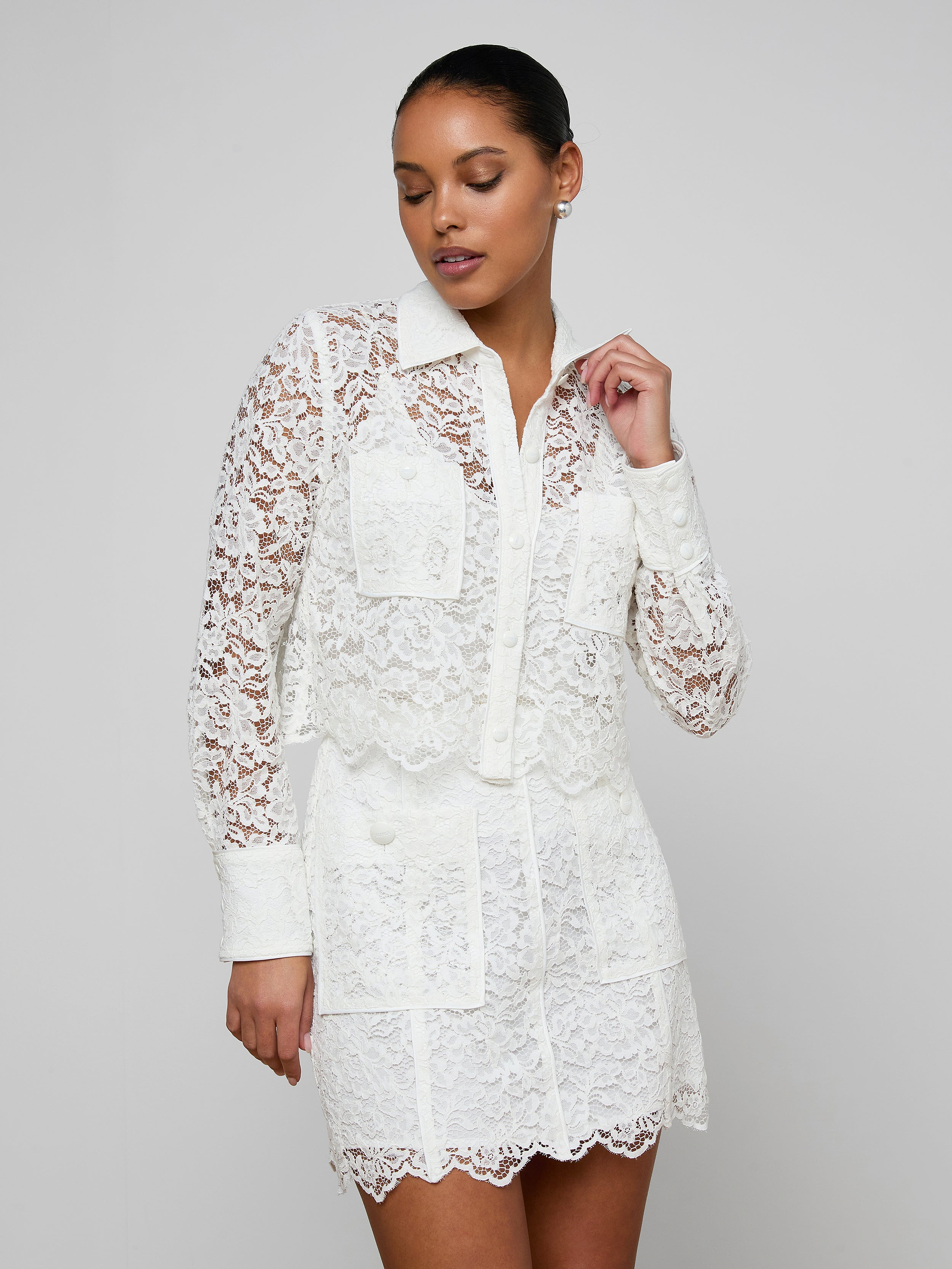 A woman wearing the Roelle Lace Blouse—a white, long-sleeve, floral lace top with patch pockets—stands against a plain light background, looking to the side with one hand raised near her face.